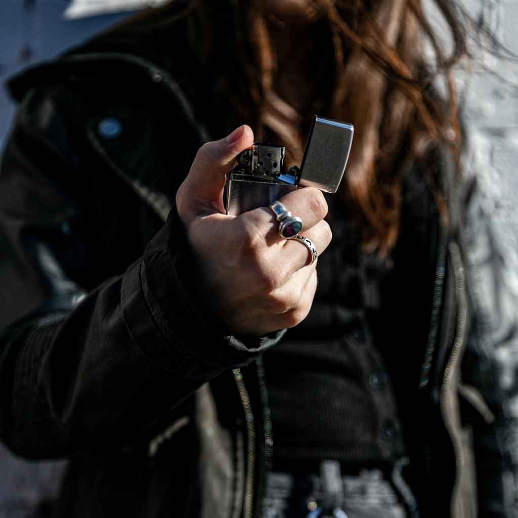 A person with long hair holds a silver lighter, wearing multiple rings on their fingers and a dark jacket, with a blurred face in the background.