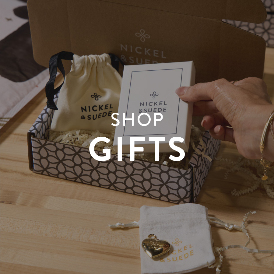 Gift box from Nickel & Suede with a drawstring bag, a gold heart pendant, and a hand holding a branded card on a wooden surface. Text reads "Shop Gifts."