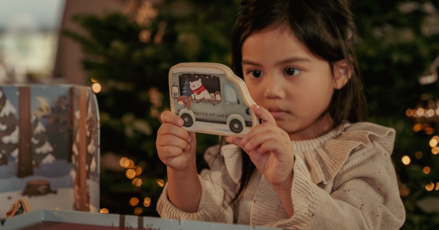 A child holds a toy van decorated with animal characters in front of a Christmas tree. Holiday-themed box is visible on the side.