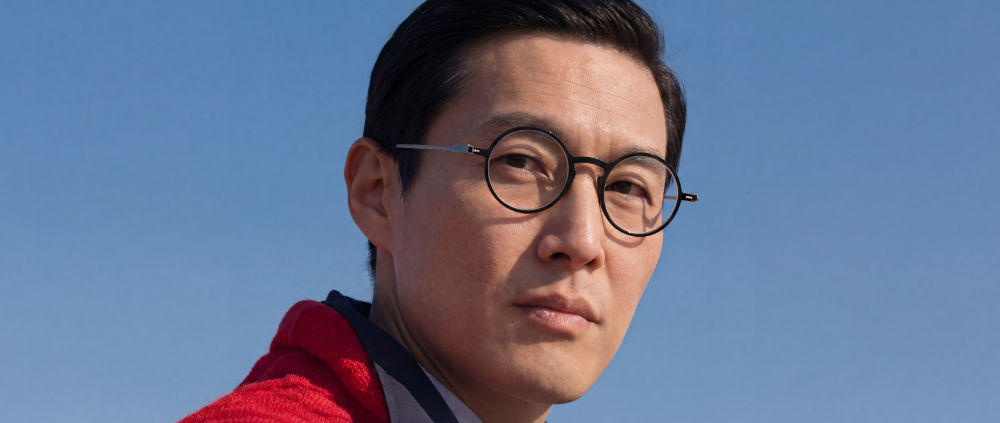 Close-up portrait of a man wearing black round reading glasses with slim metal temples, gazing upward against a clear blue sky