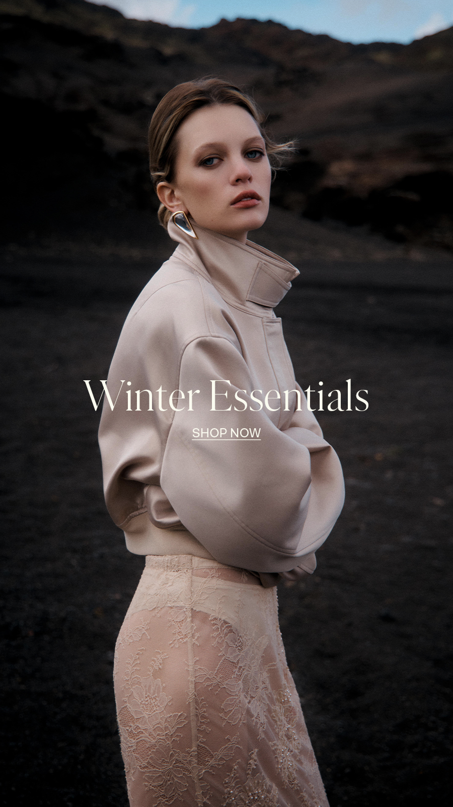 Fashion model wearing beige turtleneck jacket and lace skirt against dark rocky landscape, with "Winter Essentials Shop Now" text overlay