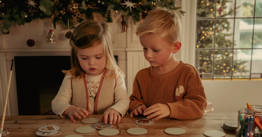 Two children seated at a table playing a card game, surrounded by Christmas decorations. A decorated tree is visible through the window.
