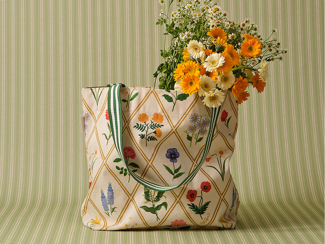 Floral-printed canvas tote bag with green striped handles, filled with yellow, orange, and white wildflowers against a green striped background.