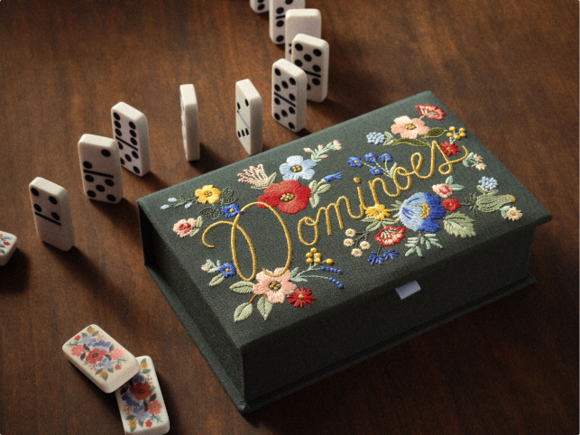 A decorative box labeled "Dominoes" with floral embroidery sits on a wooden table, surrounded by scattered domino tiles.