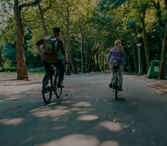 Two cyclists riding bicycles along a tree-lined park path, with a green trash bin visible on the right side.
