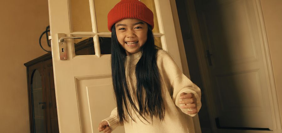 A smiling girl with long black hair wearing a red beanie hat and cream sweater, posing playfully near a white door indoors.