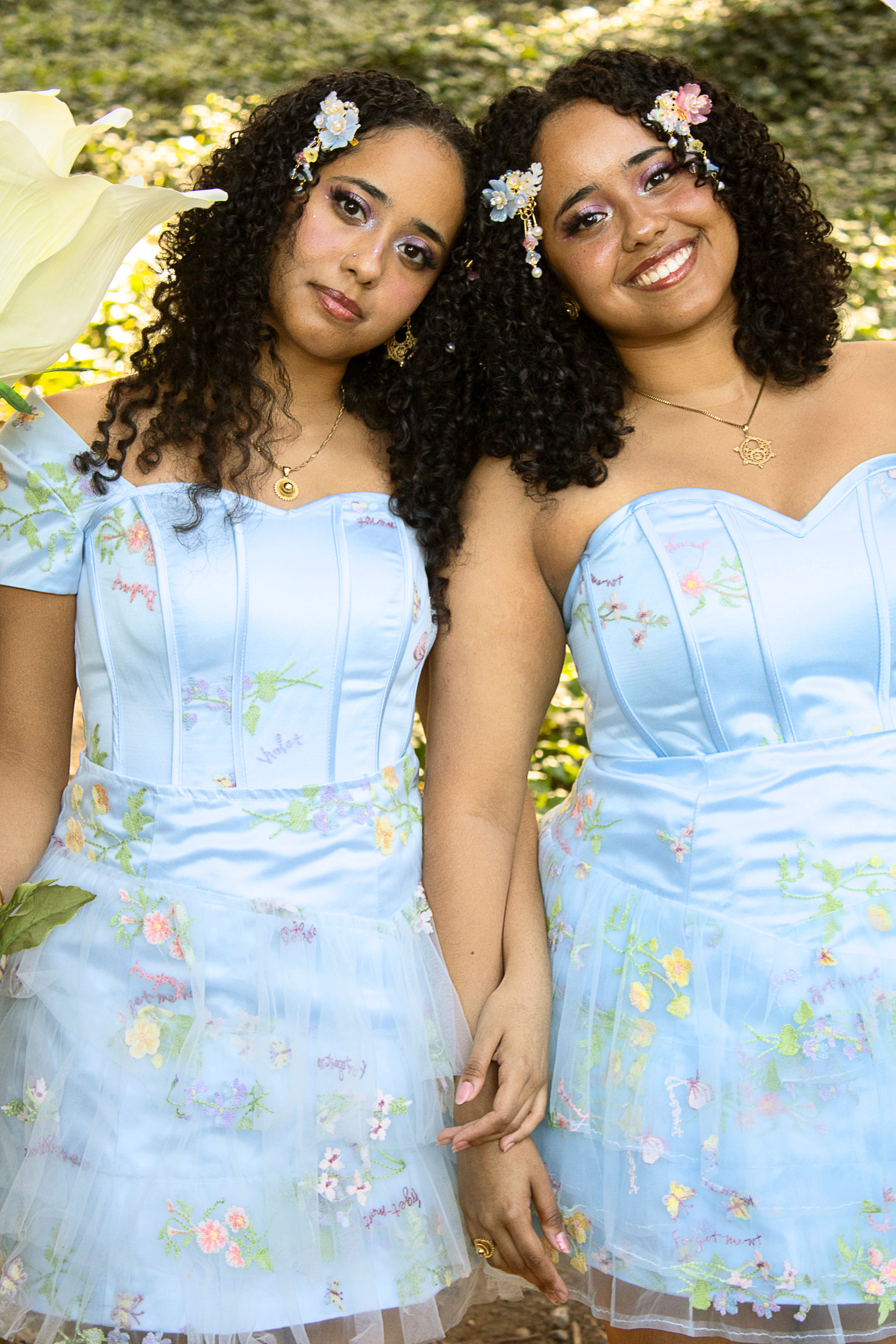 Two young women in matching light blue floral embroidered dresses with flower hair accessories, posing outdoors together.