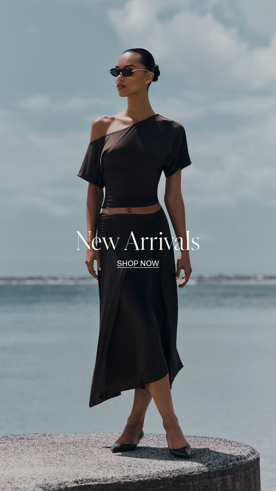 ```html
<img src="image.png" alt="Woman in brown off-shoulder dress and flats standing on a stone platform by the sea. Text reads 'New Arrivals, Shop Now'." />
```