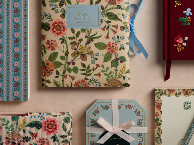 Alt text: A collection of stationery items featuring floral designs. Includes a folio with a blue ribbon, a red notebook with embroidered flowers, and other floral-patterned notebooks and paper sets arranged on a beige surface.