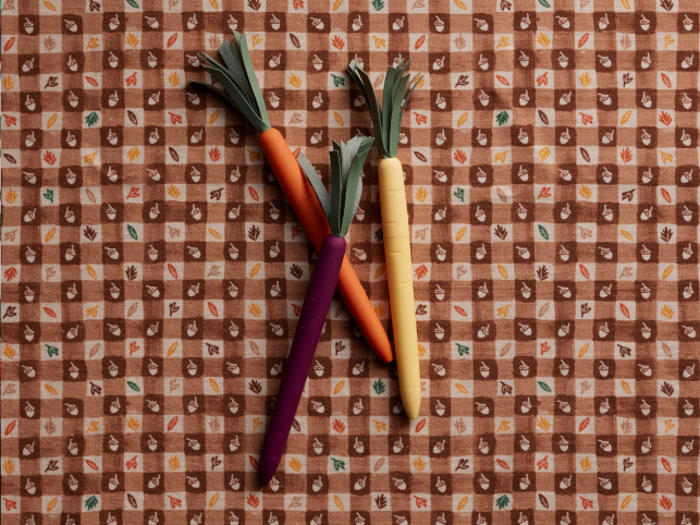 Three carrot-shaped pens in orange, purple, and yellow with green leaf tops on a brown autumn-patterned fabric background.