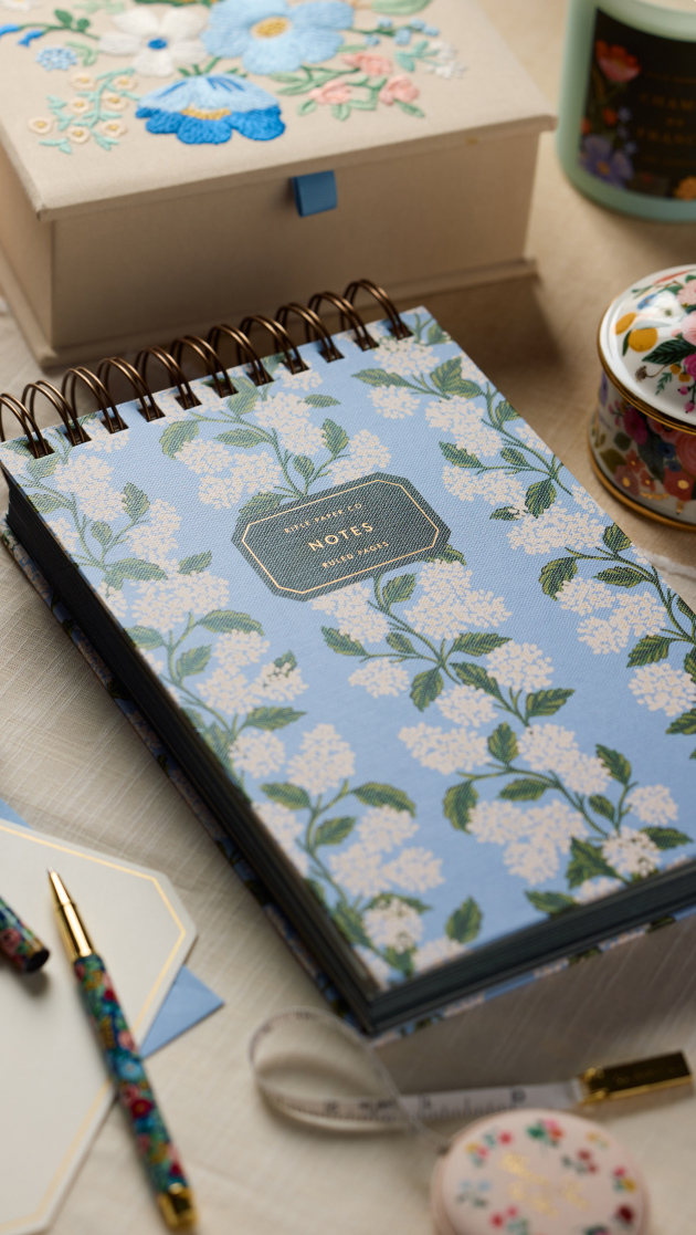 Spiral-bound notebook with floral cover design, surrounded by decorative items including a pen, embroidered box, candle, and measuring tape.