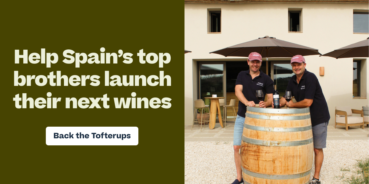 Two men in matching polo shirts and caps posing with wine bottle and glasses on a wooden barrel outside a winery building, with text "Help Spain's top brothers launch their next wines – Back the Tofterups"